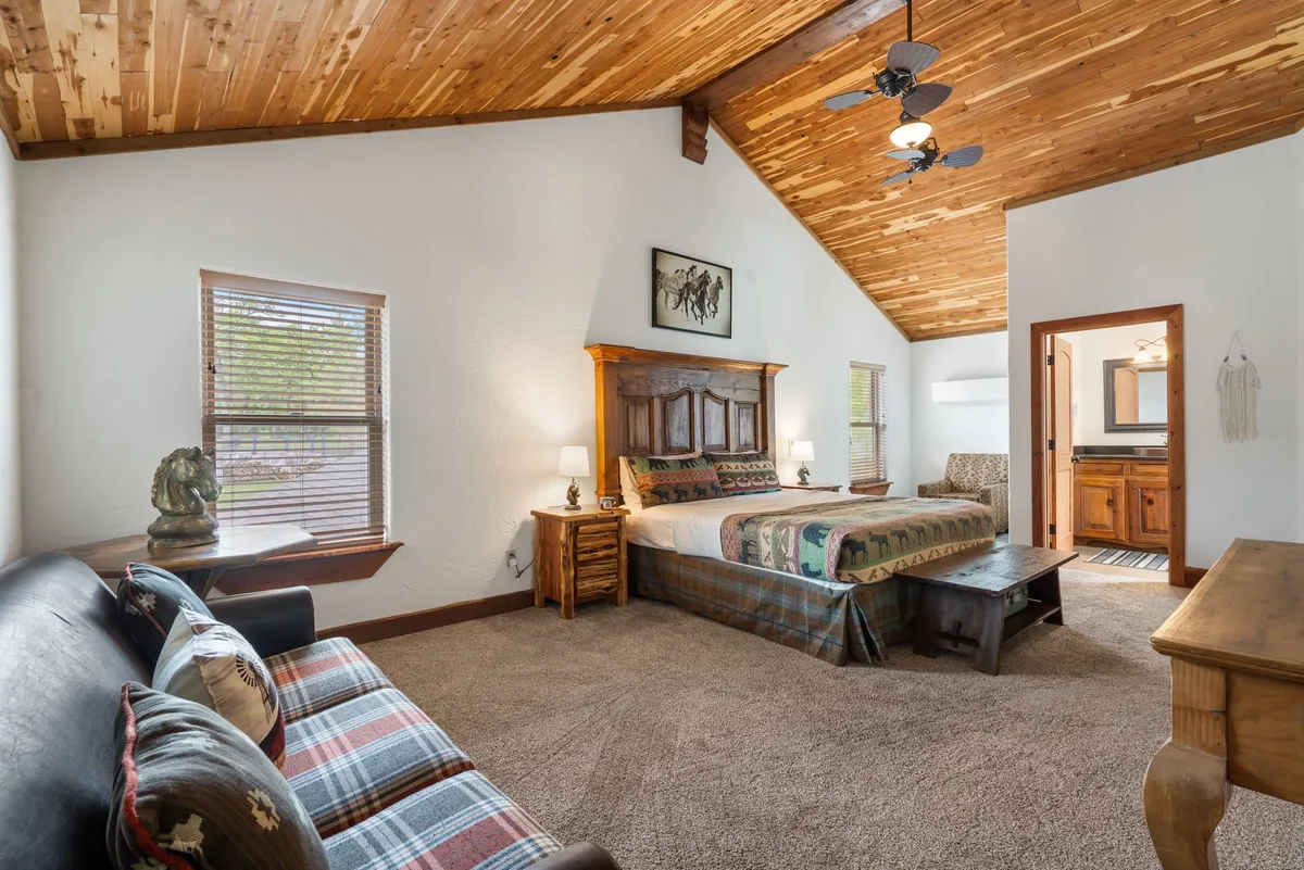A beautifully appointed bedroom with a wooden bed frame, plaid throw blanket, decorative pillows, wooden ceiling fan, and rustic artwork adorning the wall. Natural light filters in, highlighting the warm textures and cozy ambiance.