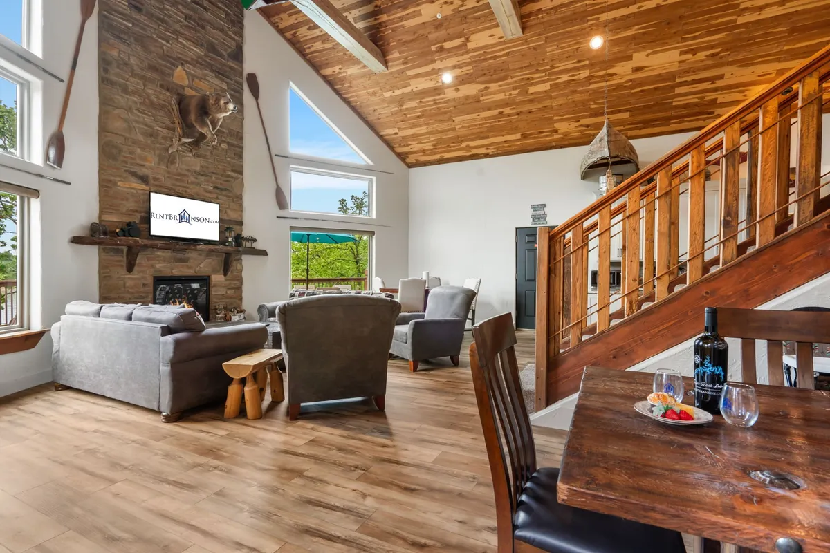 A welcoming living and dining space with a stone fireplace, comfortable seating, a large window framing scenic views, and a dining table set with wine and snacks. The room is bathed in natural light, and the setup encourages both relaxed lounging and shared meals.