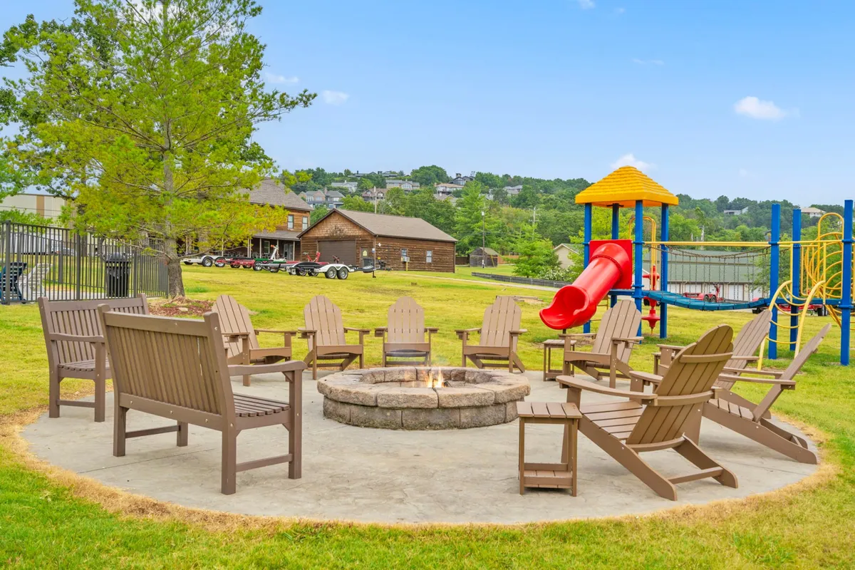 A friendly, communal gathering area featuring a fire pit surrounded by adirondack chairs, playground equipment for children, and green grass shaded by trees. The area is full of potential for family picnics, dog-friendly play, and socializing in the fresh air.