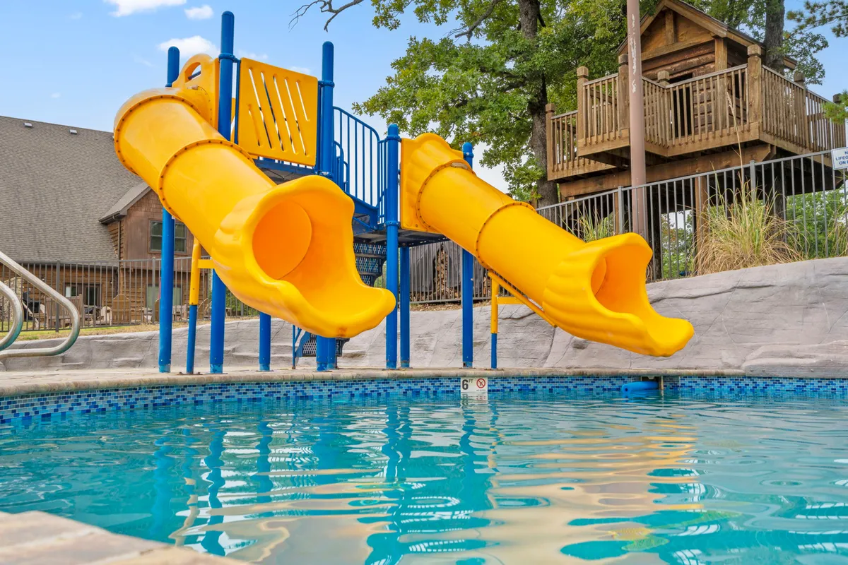 A joyful exterior pool scene featuring yellow water slides, a sparkling swimming pool, and a wooden playhouse. The area is lively with families swimming, children playing on slides, and people relaxing by the poolside.