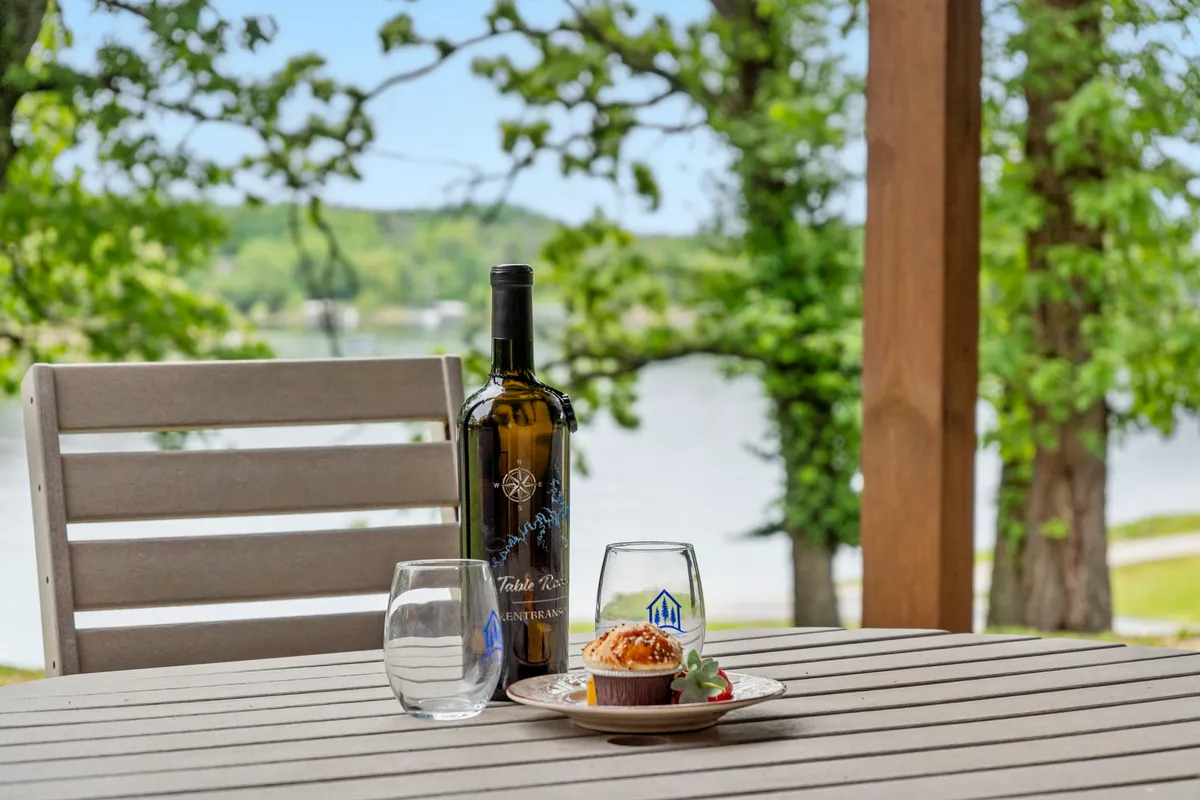 An exterior image of a beautifully set table on a lakeside deck, complete with a wine bottle, wine glass, and an elegant dessert. The table overlooks the serene water, capturing the mood of relaxed outdoor dining and celebration with friends.