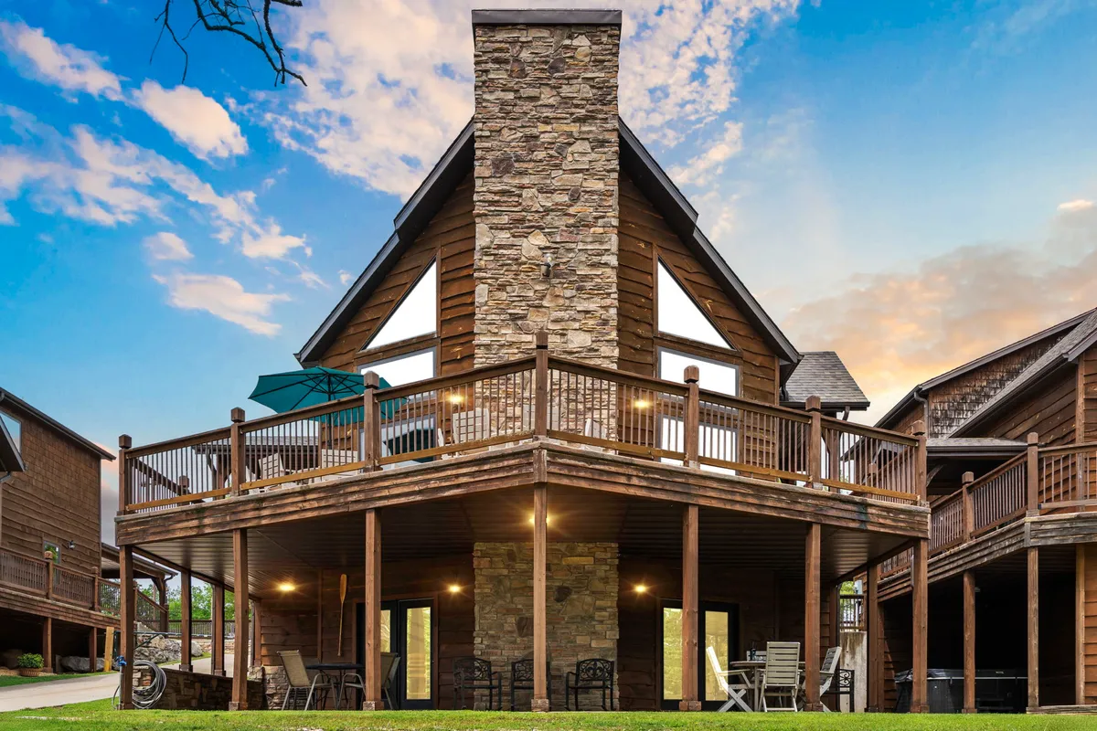 The image showcases the exterior of a stunning lakefront home at Branson Shores Resort, featuring a classic stone chimney, expansive deck with ample outdoor seating, and large windows overlooking Table Rock Lake. The scene evokes a sense of relaxation and togetherness, with visible spaces for outdoor dining, gathering with friends, and enjoying serene sunset views.