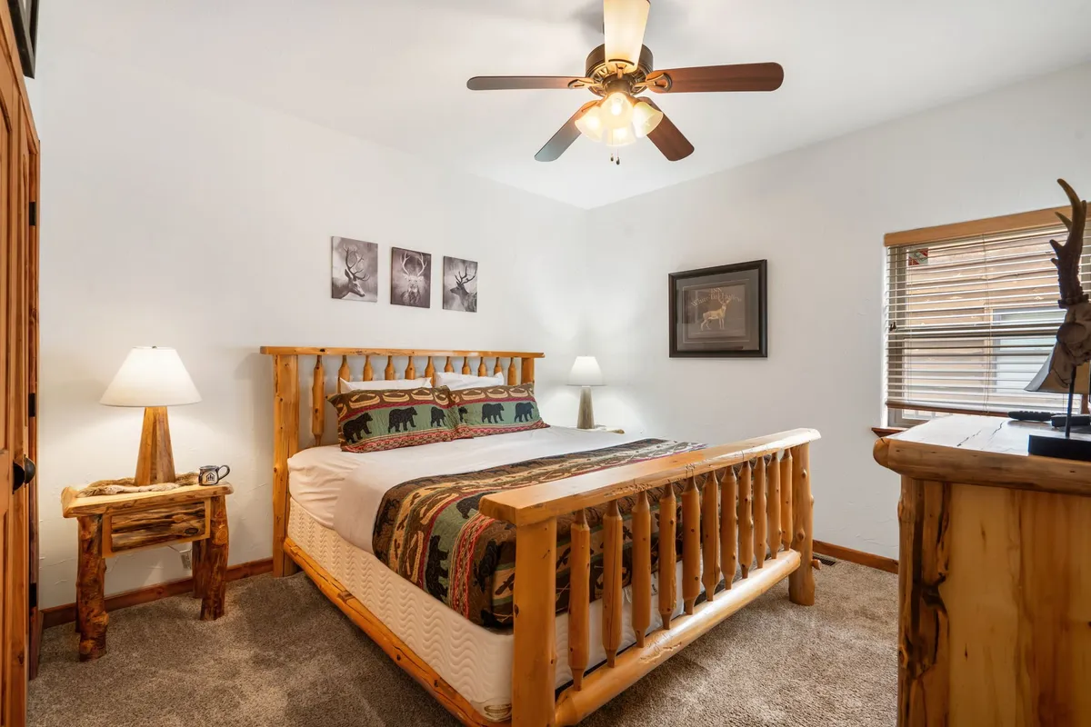 A tranquil bedroom with a log bed frame, bear-themed bedding, wooden side table, ceiling fan with light, and framed nature-inspired artwork. Daylight streams in, creating a peaceful and inviting environment.