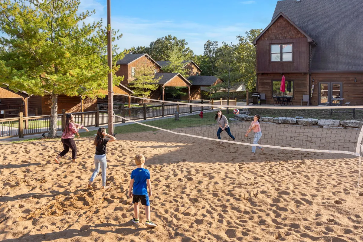 An exterior scene featuring a sand volleyball court where both children and adults are actively playing volleyball. The court is surrounded by lush trees and several resort cabins, evoking a sense of lively outdoor recreation and group activity. The mood is energetic and joyful, capturing the essence of friendly competition and togetherness.