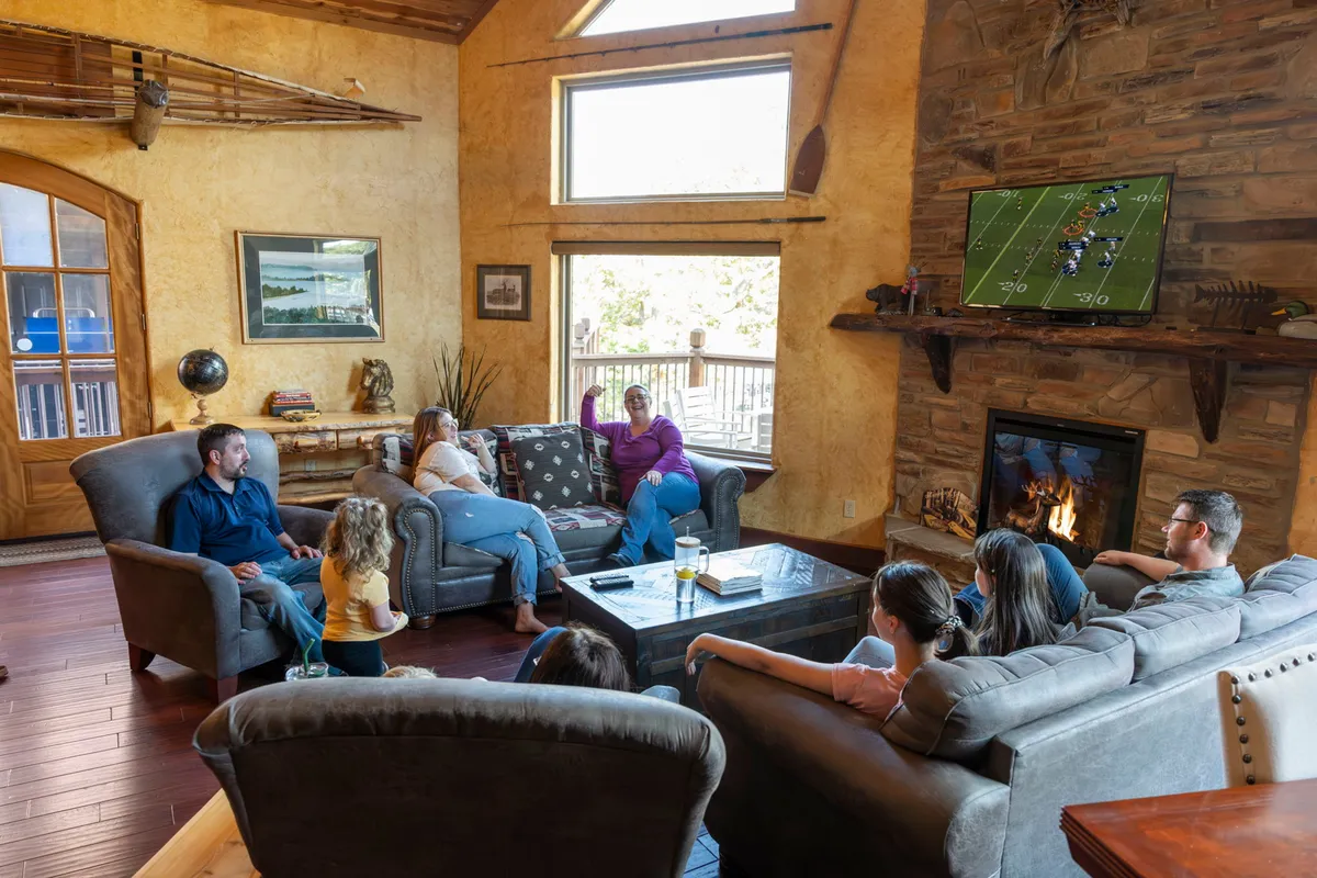 A cozy lower-level lounge featuring a large window that fills the space with natural light. The fireplace is the focal point, with a TV above showing a sports game. A family is gathered on comfortable sofas, enjoying snacks and drinks while watching together.