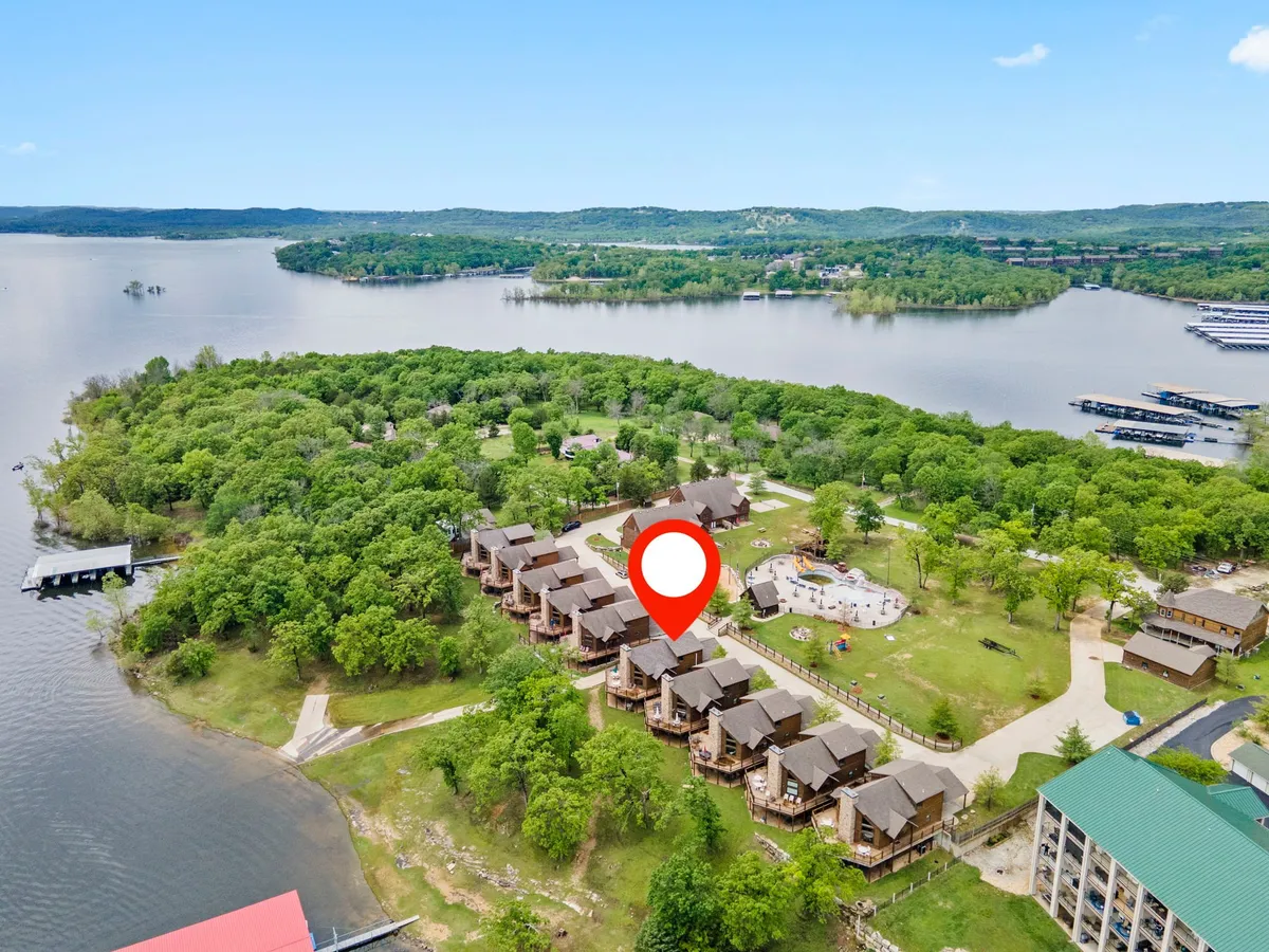 This image captures a tranquil lakeside scene featuring cabins nestled among greenery, a visible playground, and a dock extending into the calm water. The layout invites the viewer to imagine families and their dogs enjoying the open space, exploring the shore, or playing on the playground together.