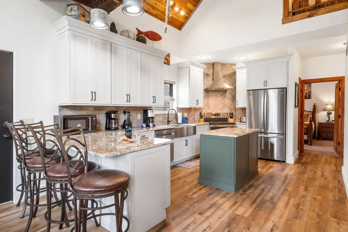 A spacious kitchen with a central island features granite countertops, stainless steel appliances, bar stools for casual seating, and a warm, wooden ceiling. The layout is ideal for cooking together, chatting over breakfast, or preparing group meals, with ample space for everyone to participate.